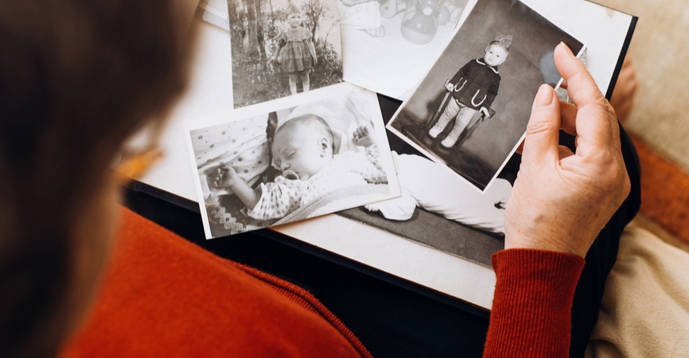 A photo album is viewed from over an elderly woman’s shoulder. She is sorting through a series of old black and white photographs.